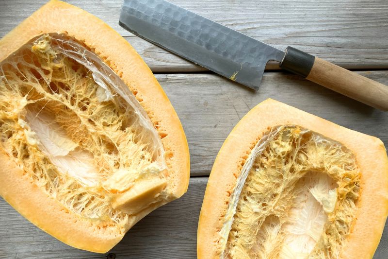 A spaghetti squash cut in half against a wooden background with a sharp Japanese chef's knife on top