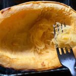 A fork twirling strands of spaghetti squash in front of a cooked half a spaghetti squash