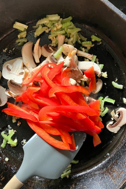 Chopped celery, mushrooms, garlic, and bell peppers with a grey silicone spatula and cast iron skillet